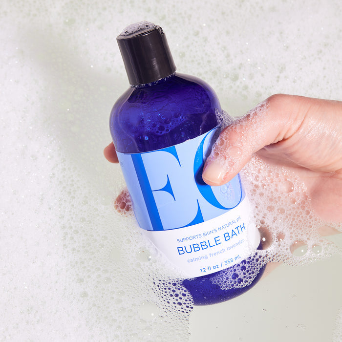 A hand gently holds a blue EO Products Bubble Bath bottle, infused with calming French lavender. Amidst foamy bubbles, this serene bath setting offers a tranquil escape with its soothing scent.