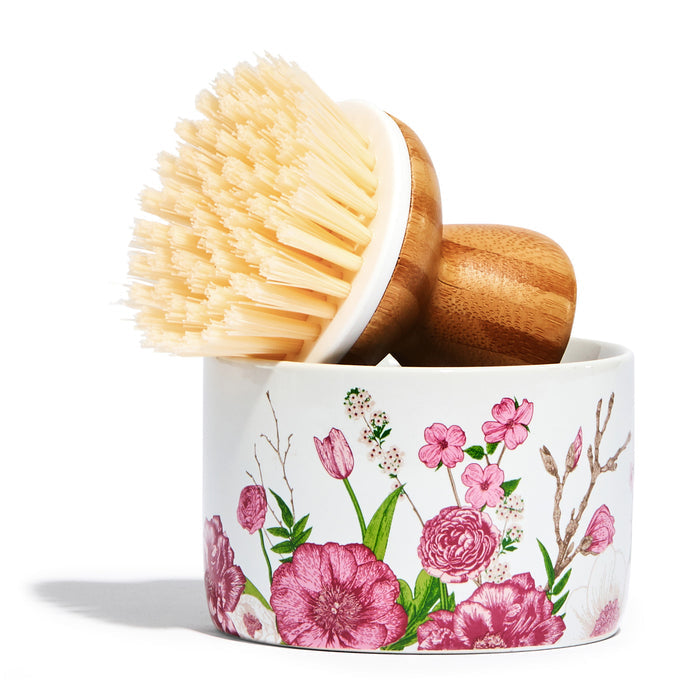 A Grove Co. Bubble Up Dish Soap Dispenser & Brush Set features a dish scrub brush with beige bristles and a wooden handle inside a ceramic holder adorned with colorful pink and red floral designs, including various flowers and green leaves.