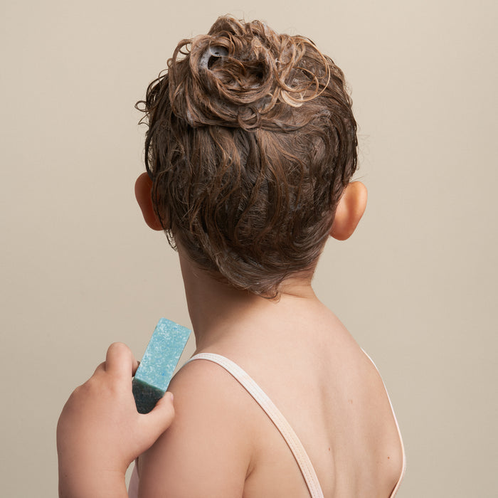 A child with wet, sudsy hair holds a Bubble-Yum Body Bar from NOT!CE Hair Co against a neutral beige background. Wearing a light top, they showcase the joy of plastic-free hair care using vegan and cruelty-free products for fun bath time.