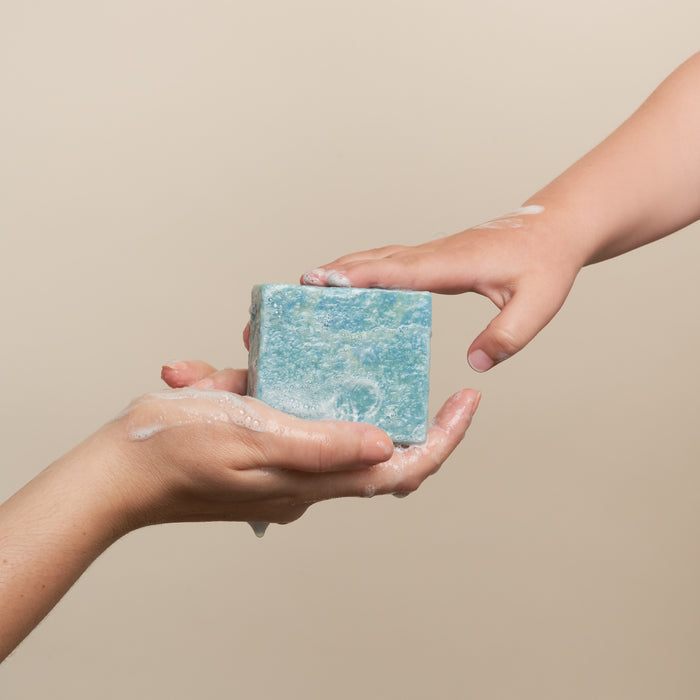 Two hands, covered in soap suds, exchange a large blue Bubble-Yum Body Bar from NOT!CE Hair Co against a neutral background, emphasizing its vegan and cruelty-free qualities.