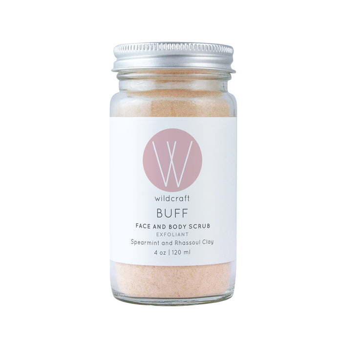 A 4 oz (120 ml) glass jar of Wildcraft Buff Face and Body Scrub with a metal lid features a white label with a pink W logo, describing it as an exfoliant containing spearmint and rhassoul clay.