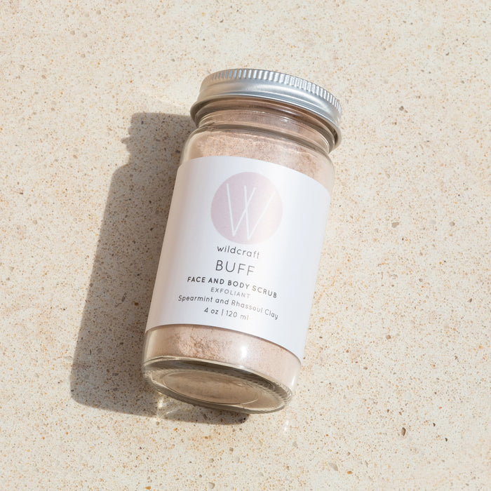 A clear jar with a silver lid containing Wildcraft Buff Face and Body Scrub, enhanced by spearmint and rhassoul clay for a natural scent, sits on a textured surface casting a soft shadow.