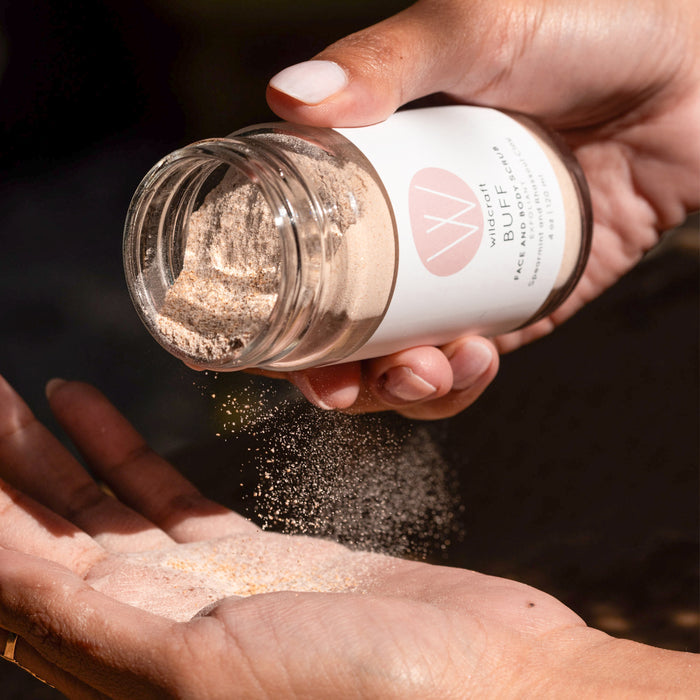 A person pours Wildcrafts Buff Face and Body Scrub into their palm. The powder, with its natural scent, catches the light as it falls, creating a soft texture against a dark background, highlighting the exfoliators action and allure.