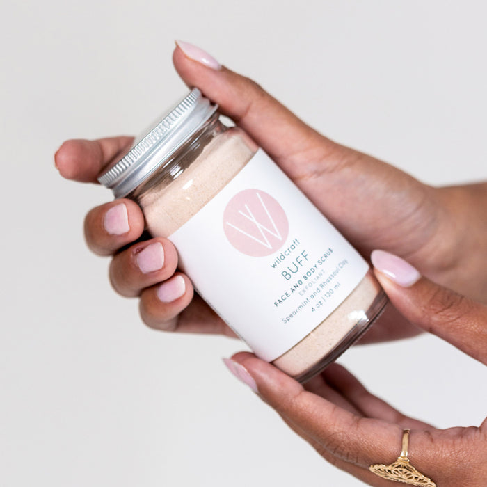 Hands gently cradle a jar of Wildcrafts Buff Face and Body Scrub, an ideal exfoliator with a natural scent. Its metal lid pairs well with the white label featuring pink accents, coordinating beautifully with the persons pale pink nails.