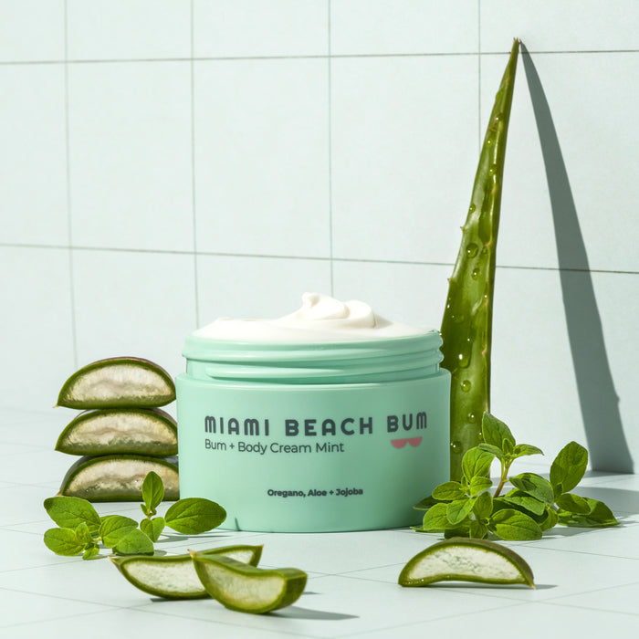 A green jar of Miami Beach Bums Bum + Body Cream in mint, with the lid off, is surrounded by aloe vera slices and leaves. The label highlights oregano, aloe, and jojoba for skin health. This is set against a tiled background.
