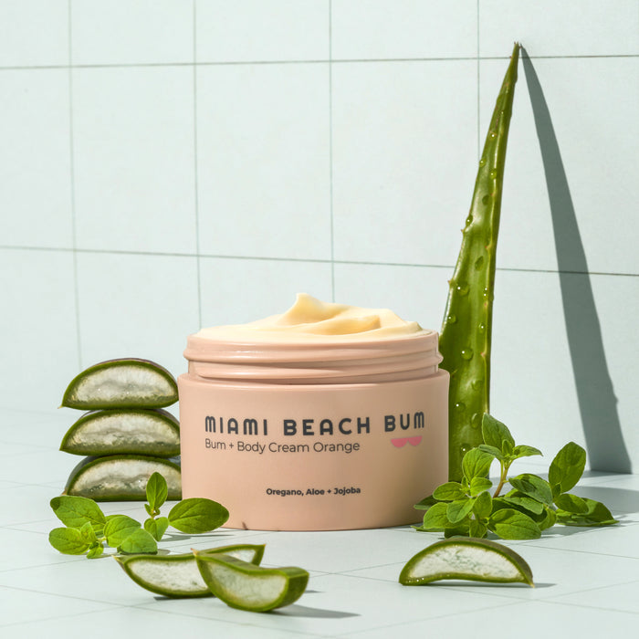 An open jar of Miami Beach Bums Bum + Body Cream showcases its creamy texture. Surrounded by aloe vera slices, an aloe leaf, and fresh green leaves, it emphasizes natural ingredients like oregano for improved skin health against a tiled background.