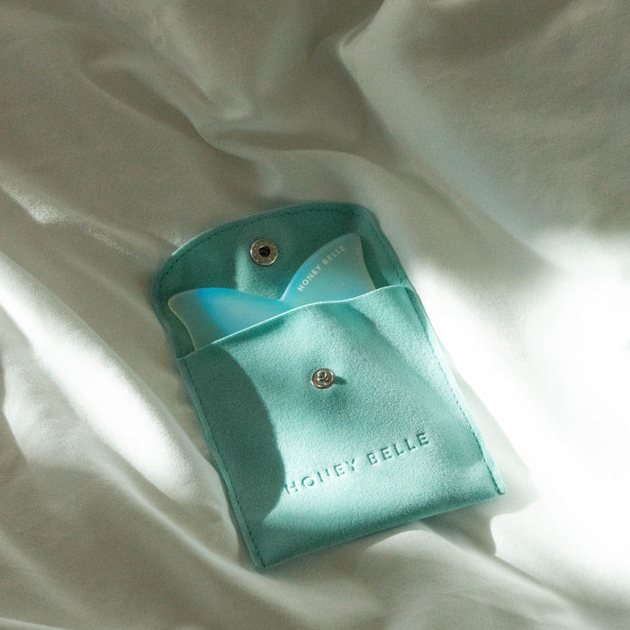 A mint green pouch marked with Honey Belle reveals the Butterfly Gua Sha tool in blue and white, designed for contouring. The open pouch lies on soft, light-colored fabric.