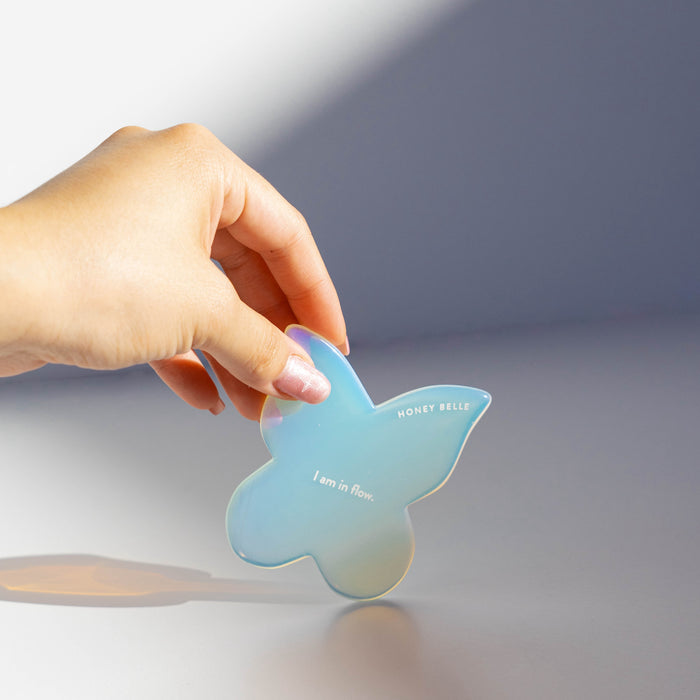 A hand holds the iridescent Butterfly Gua Sha by Honey Belle, perfect for lymphatic drainage and contouring. HONEY BELLE and I am in flow. are printed on it. A soft light casts a gentle shadow, accentuating its elegance.