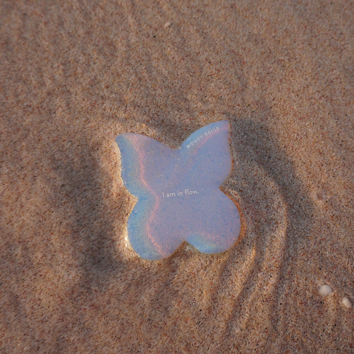 A Butterfly Gua Sha sticker by Honey Belle, featuring I am in flow, sits on wet sand, mirroring the smoothness of a Gua Sha tool. The sands contours and gentle water ripples evoke the calmness of lymphatic drainage.