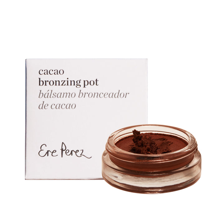 A small glass pot in a dark brown hue contains smooth, natural bronzing cream from Ere Perez. It sits alongside a white box labeled Cacao Bronzing Pot Bálsamo Bronceador de Cacao, promising an effortless sun-kissed glow.