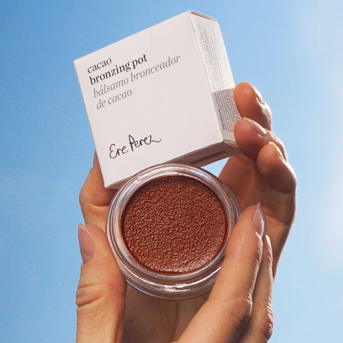 Hands gently hold a small glass container labeled Cacao Bronzing Pot by Ere Perez, containing natural bronzing cream. Set against a clear blue sky, the elegant white packaging box enhances the allure of this face bronzer.
