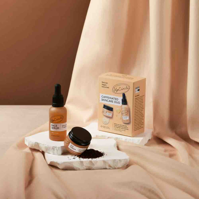 On a beige fabric background, UpCircles Caffeinated Skincare Duo is displayed with an organic face serum dropper, eye cream jar, and matching box. Upcycled coffee oil-infused grounds are scattered on a white tray.