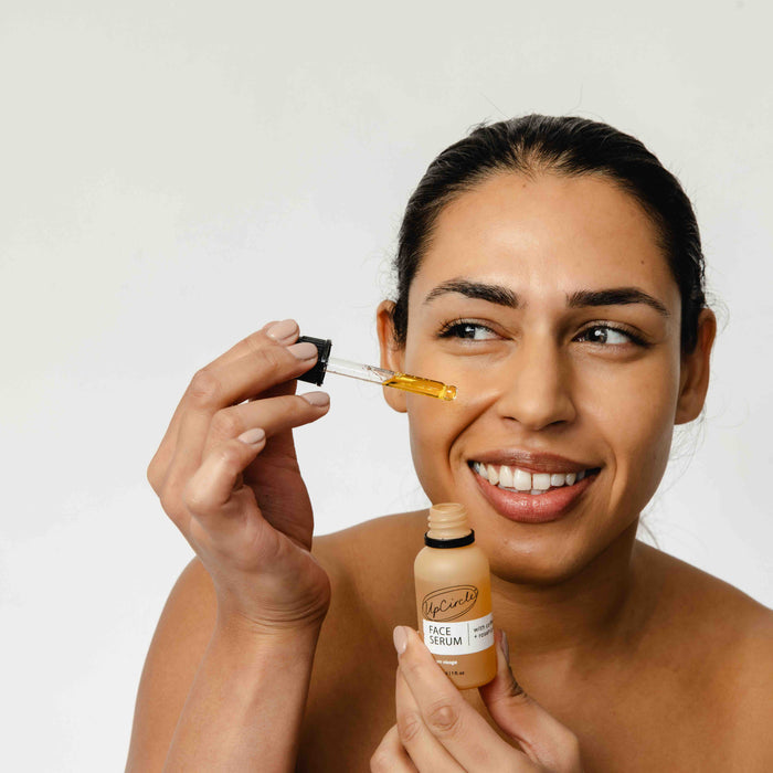 A woman smiles while applying UpCircles Caffeinated Skincare Duo with a dropper. Holding the bottle in her left hand, she applies the nourishing serum, infused with upcycled coffee oil, to her right cheek against an off-white background that highlights her radiant glow.