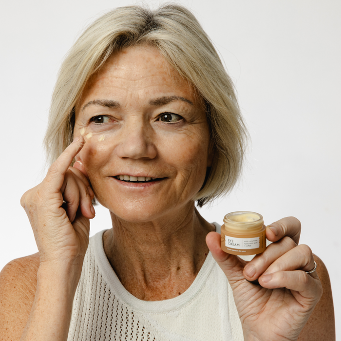 A smiling older woman with short blonde hair applies UpCircles Caffeinated Skincare Duo, enriched with upcycled coffee oil, to her eye area. Shes dressed in a white sleeveless top against a plain white background, exuding youthful energy.