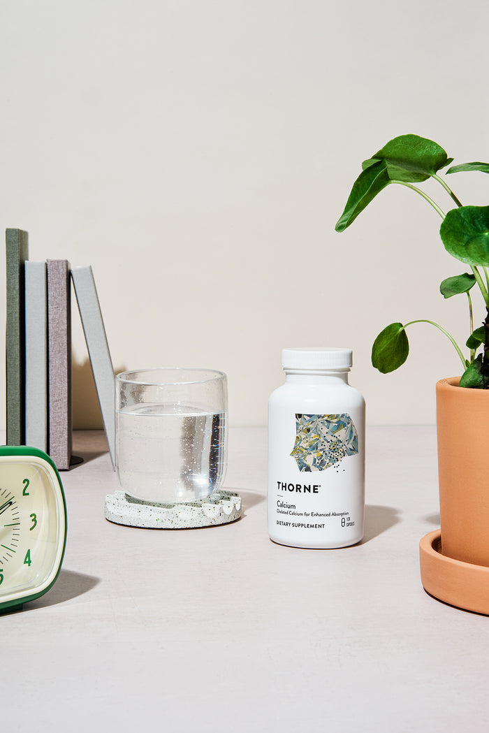 A white Thorne bottle labeled Calcium sits on a table with a glass of water, potted plant, and books. A white clock with green trim adds to the calm setting—ideal for peri- and postmenopausal women seeking enhanced absorption in their calcium supplement.