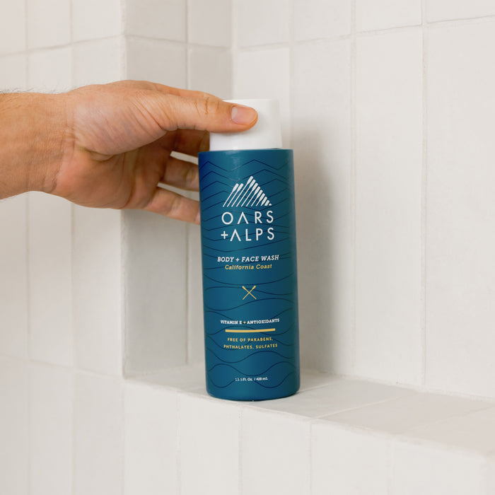 A hand reaches for a blue bottle of Oars + Alps Cali Coast Body Wash, enriched with Vitamin E and free of parabens, phthalates, and sulfates, resting on a white tiled shower shelf.