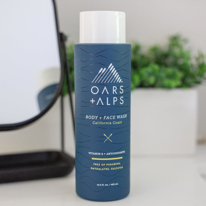 A blue bottle of Oars + Alps Cali Coast Body Wash sits on a white surface with a blurred plant behind it. The label notes Vitamin E, its California Coast scent, and that it’s free of parabens, phthalates, and sulfates.