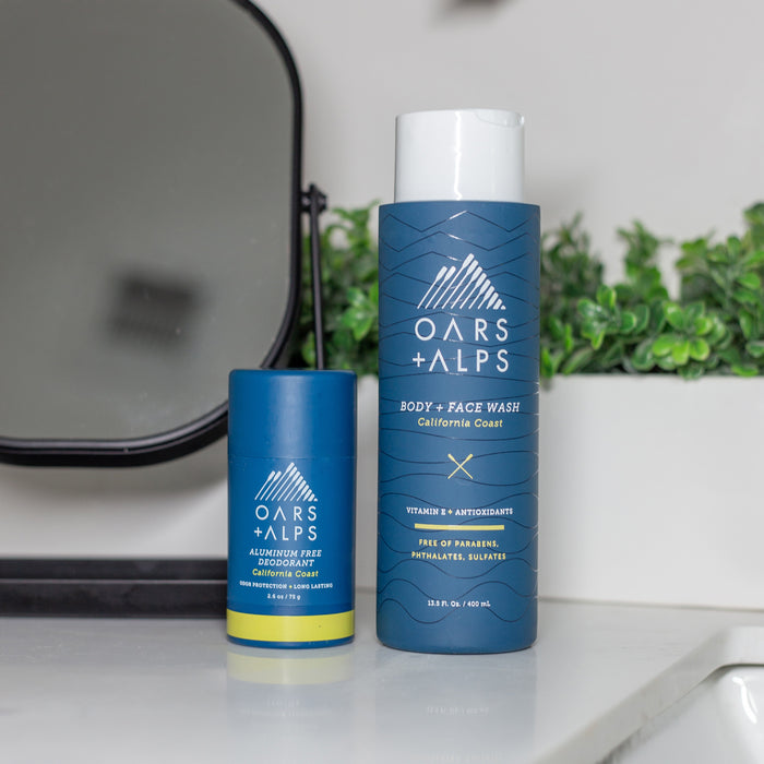 Two Oars + Alps products on a bathroom counter: a blue tube of aluminum-free deodorant and a larger blue bottle of Cali Coast Body Wash. A mirror and green plant are in the background.