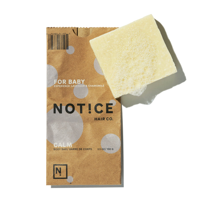 A brown paper package with white polka dots labeled NOT!CE Hair Co. and For Baby: Lavender & Chamomile caters to sensitive skin, accompanied by a Calm Body Bar—a square, pale yellow, textured soap infused with soothing lavender essential oil.