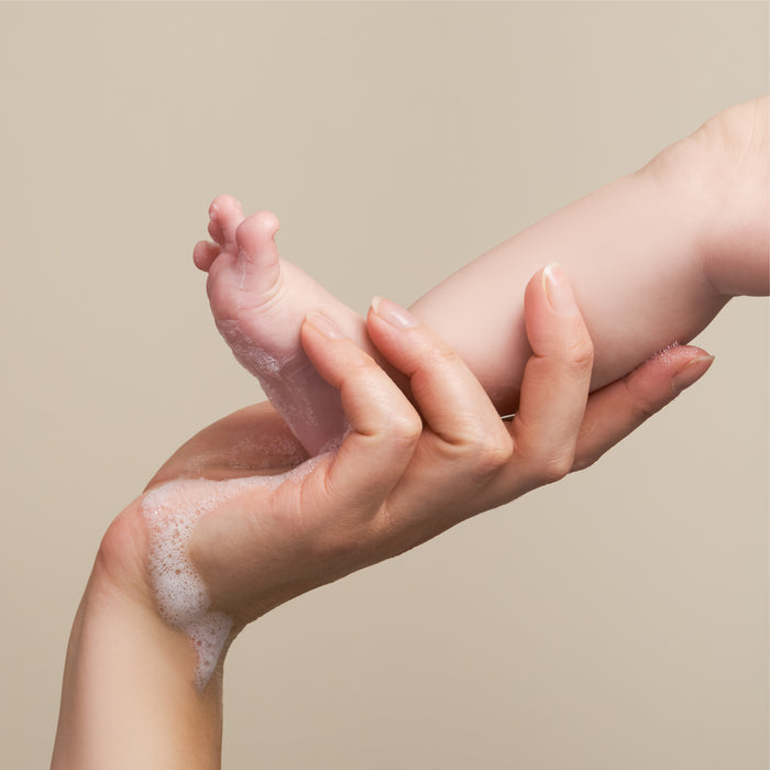 An adults soapy hand gently holds a babys foot against a neutral background. The NOT!CE Hair Co Calm Body Bar, enriched with sea salt, keeps the babys sensitive skin soft and cared for as soap suds tumble during washing.