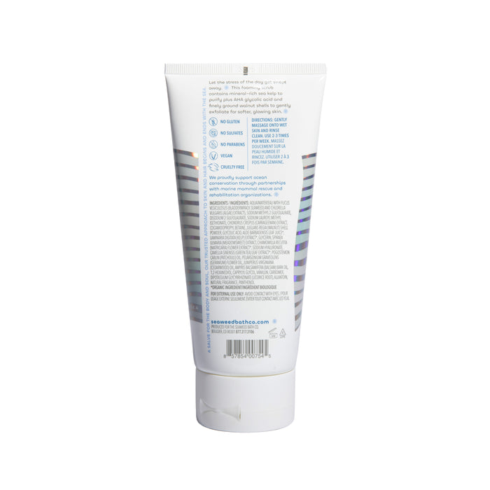 The image shows the back of The Seaweed Bath Co’s Calm Body Scrub, a white plastic tube. It lists ingredients like AHA glycolic acid and sea kelp extract to exfoliate and nourish your skin. Brand details, a barcode, and a website URL are also visible at the bottom.