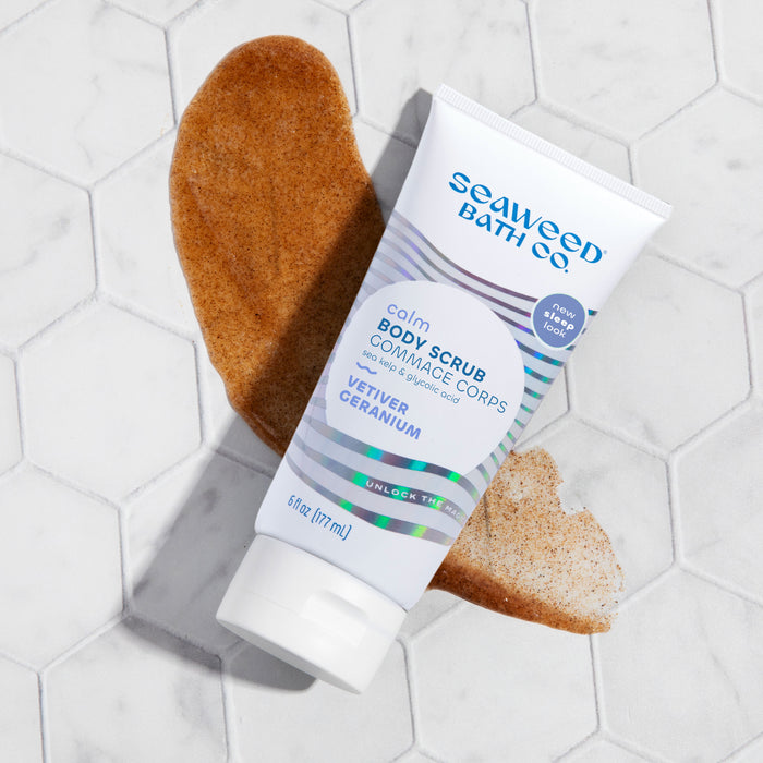 A tube of The Seaweed Bath Co Calm Body Scrub with a new look rests on white hexagonal tiles, promising to exfoliate with sea kelp and AHA glycolic acid. Two smears of the vegan scrub highlight its blend.