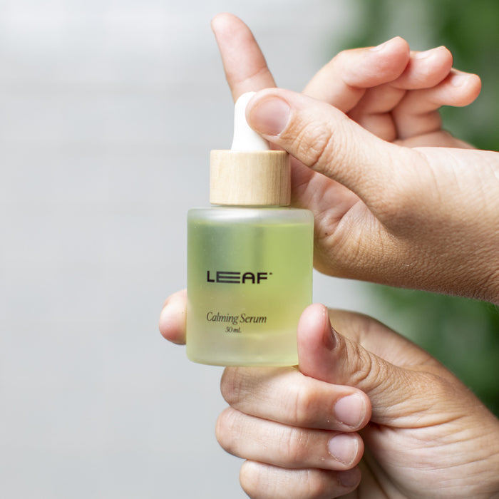 Close-up of hands holding a glass bottle labeled Calming Serum w/ Dropper by Leaf Shave. It has a wooden cap and contains light green, antioxidant-rich liquid. The softly blurred background enhances its soothing appearance.