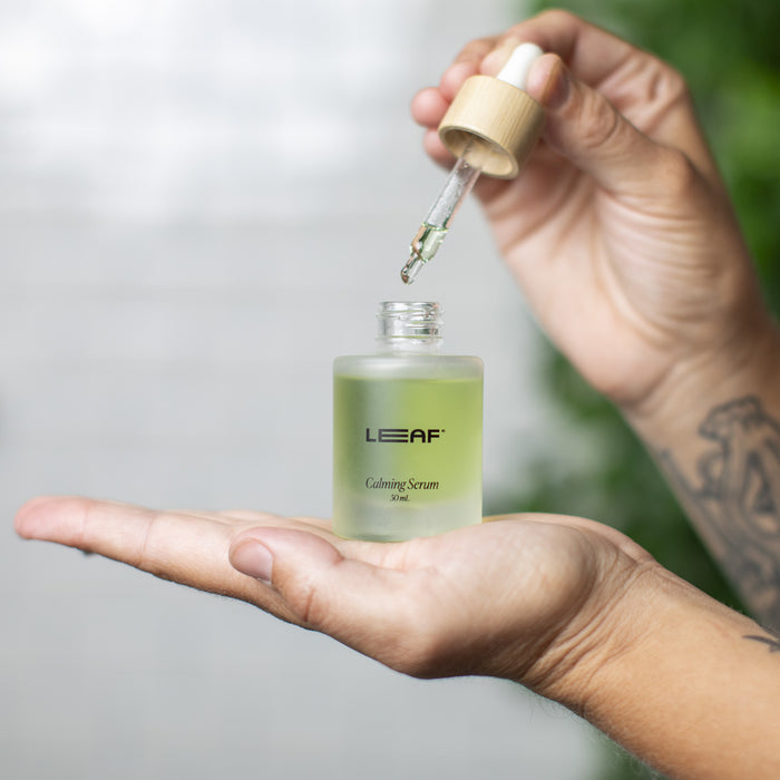 Holding a small bottle of Leaf Shaves Calming Serum w/ Dropper, a person uses the dropper with the other hand. This vegan serum, packed with skin-soothing antioxidants, is set against a backdrop of blurred plants.