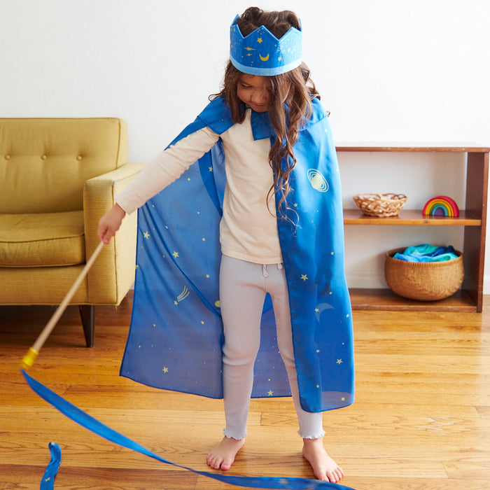 A child in a Sarahs Silks cape and blue starry crown stands on wooden flooring, holding a wand. Nearby are a yellow chair and a shelf with a basket and rainbow toy, creating the perfect setting for imaginative dress-up play.