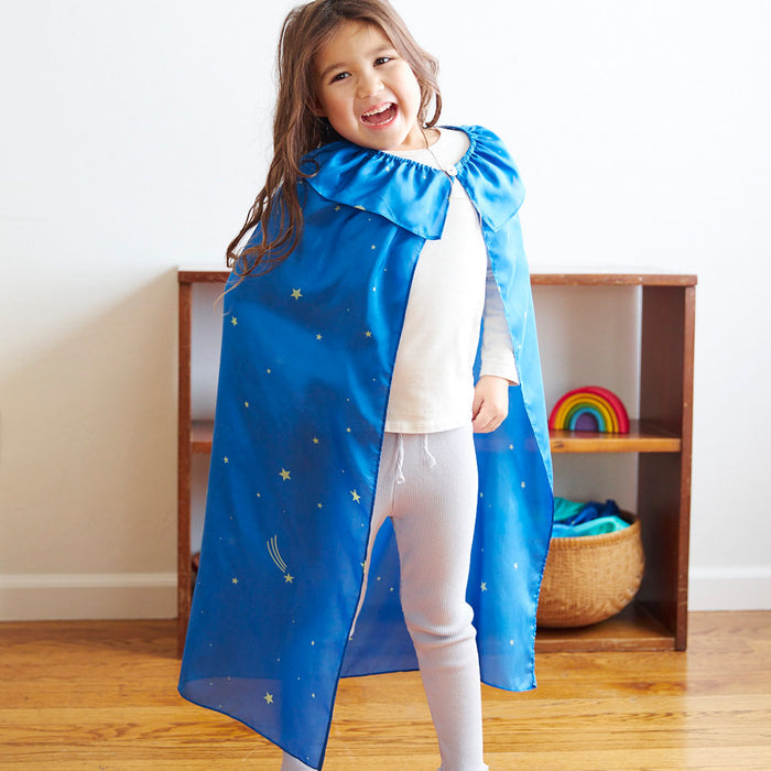 A smiling child with long brown hair wears a white shirt, gray leggings, and a Sarahs Silks cape adorned with stars and a shooting star, ideal for dress-up. In the background are a small shelf with a rainbow decoration and a woven basket.