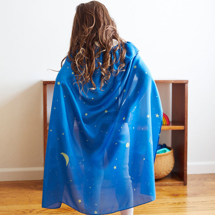 A child with long brown hair stands with their back to the camera, wearing a silk cape from Sarahs Silks, decorated with yellow stars and crescent moons. In a room with wooden floors, they engage in dress-up play near a shelf filled with colored balls and a basket.