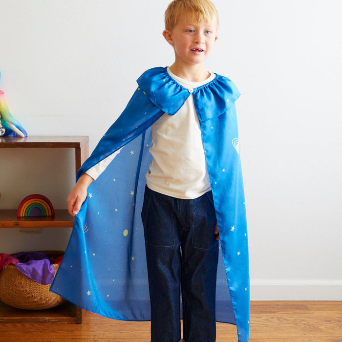 A young child stands smiling in a room, engaging in dress-up play while wearing a Cape by Sarahs Silks, embellished with stars over a white shirt and jeans. Nearby, a shelf displays toys, including a small rainbow decoration.