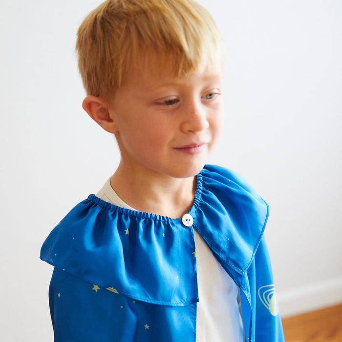 A young blond boy wears a Sarahs Silks blue cape decorated with stars and planets. While engaging in dress-up play, he smiles slightly and looks to his right. The silk cape, fastened with a button, is set against a simple white background.