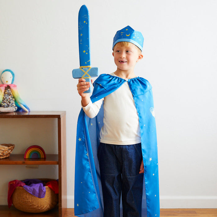 A child stands in a room wearing Sarahs Silks blue star-patterned silk cape and crown, holding a toy sword. In the background, a shelf displays a colorful doll and rainbow toy. The scene radiates joy and dramatic play.