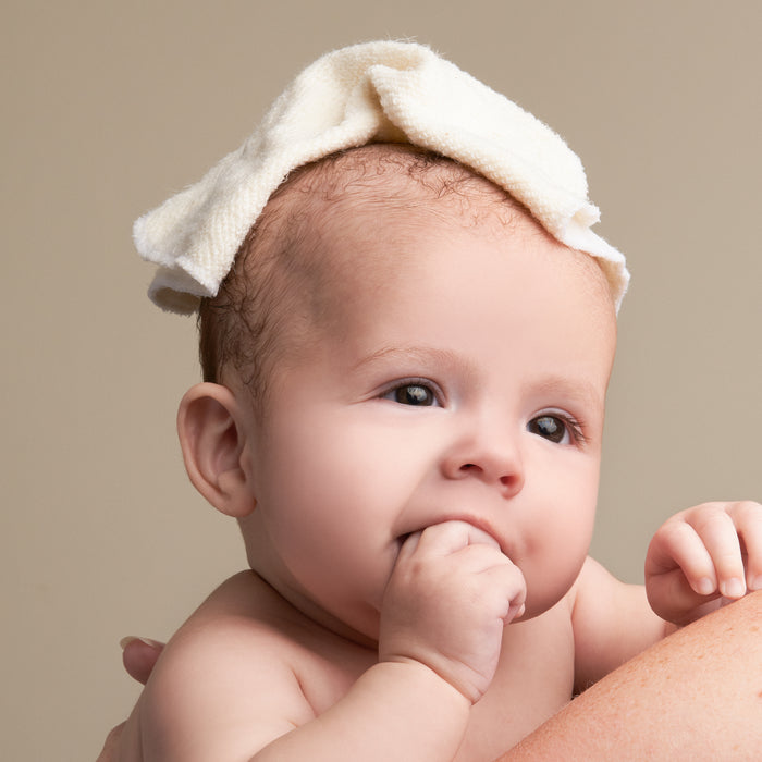 A baby with a thoughtful expression wears a Caress Reusable Cloth by NOT!CE Hair Co made of hypoallergenic, handmade organic bamboo. The baby is sucking their thumb against a neutral beige background, highlighting their smooth complexion and tender features.