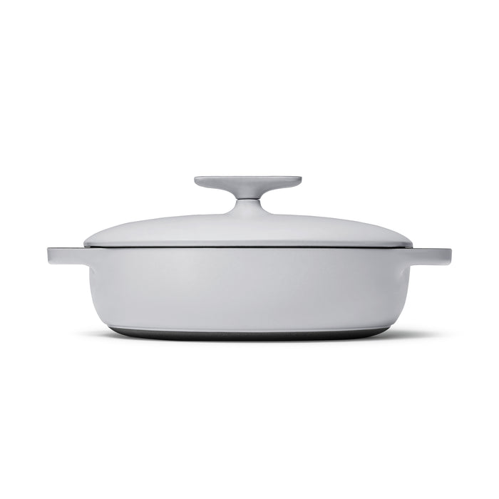 The Caraway 3.5 qt Cast Iron Braiser with Lid is shown from the side against a white background. This round, white Dutch oven features two handles, superior heat retention with a matte finish, and a durable enamel coating, complete with a lid knob.
