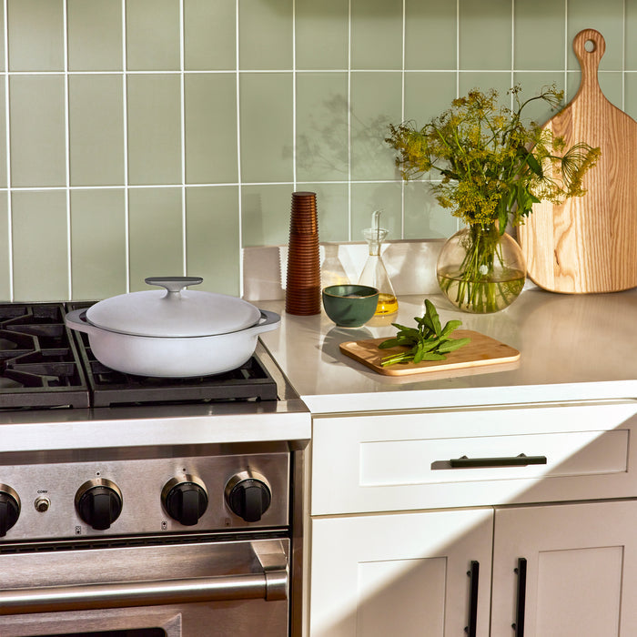 In a modern kitchen, a Caraway Cast Iron Braiser with Lid (3.5 qt), renowned for superb heat retention, sits on the stovetop. A cutting board with herbs, flowers in a vase, and oil bottles adorn the countertop against a green tiled backsplash while natural light floods the space.