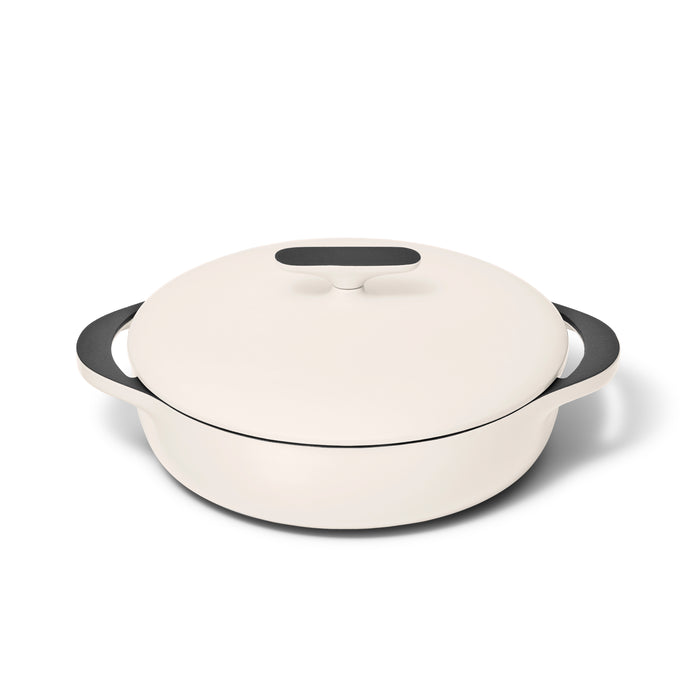 The Caraway Cast Iron Braiser with Lid, 3.5 qt, features a cream-colored round design, excellent heat retention, two black handles, and a black knob on the lid. It sits on a white background, combining style and functionality for any kitchen.
