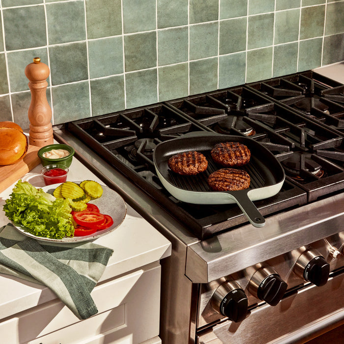 A kitchen scene showcases the 10.5 Caraway Cast Iron Grill Pan on the stove, sizzling with burger patties. Nearby, a counter holds fresh toppings: sliced tomatoes, pickles, lettuce, and small bowls of ketchup and mayo. A pepper grinder and kitchen towel complete this culinary setup.
