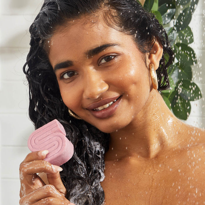 A person with wet, color-treated hair smiles while holding a pink, S-shaped Castor Oil Nourishing Shampoo Bar by Kitsch. Water droplets glisten on their skin as they stand among green plants against a white tiled backdrop.