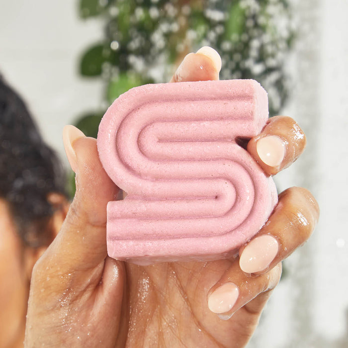 A person holds Kitschs Castor Oil Nourishing Shampoo Bar, shaped like an S and colored pink, in their wet hands. The backdrop of green leaves with a water blur evokes a refreshing shower scene ideal for soothing scalp irritation.