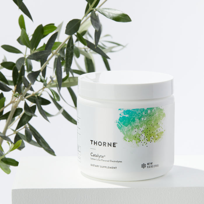 A white container of Thornes Catalyte, a dietary supplement for electrolyte restoration, sits on a white surface. The container showcases a colorful abstract design. In the background, green leaves suggest natural endurance recovery.