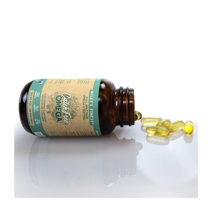 A brown bottle labeled Wileys Finest Catchfree Omega-3 lies on its side, spilling yellow Omega-3 capsules. The white background emphasizes the labels graphics and text detailing nutritional benefits and vegan DHA content.