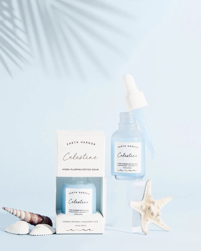 Skincare items, featuring a dropper bottle and Earth Harbor CELESTINE Hydra-Peptide Replenishing Serum box, are on a light blue surface. Seashells and a starfish evoke celestial radiance, while palm leaf shadows enhance the vegan, cruelty-free display.