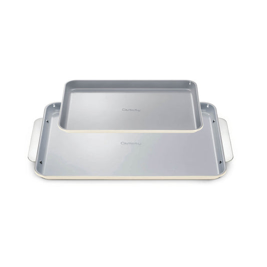The Caraway Ceramic Baking Sheet Duo features two overlapping sheets with white edges and light gray, naturally non-stick surfaces, complete with extended side handles for easy grip. They boast a non-toxic design perfect for healthy baking and are set against a white background.
