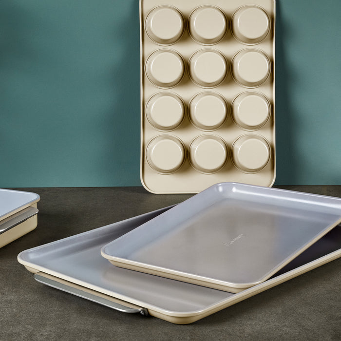 Caraways Ceramic Baking Sheet Duo and muffin tray, with 12 round molds, are elegantly displayed on a dark surface with a teal backdrop. These naturally non-stick and non-toxic items are perfect for healthy baking.