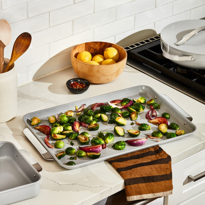 The Caraway Ceramic Baking Sheet Duo, praised for being non-toxic and naturally non-stick, holds roasted Brussels sprouts and red onion slices on the kitchen counter. Nearby are a wooden bowl with lemons, a white pot of wooden utensils, a striped towel, and a small bowl with red pepper flakes.