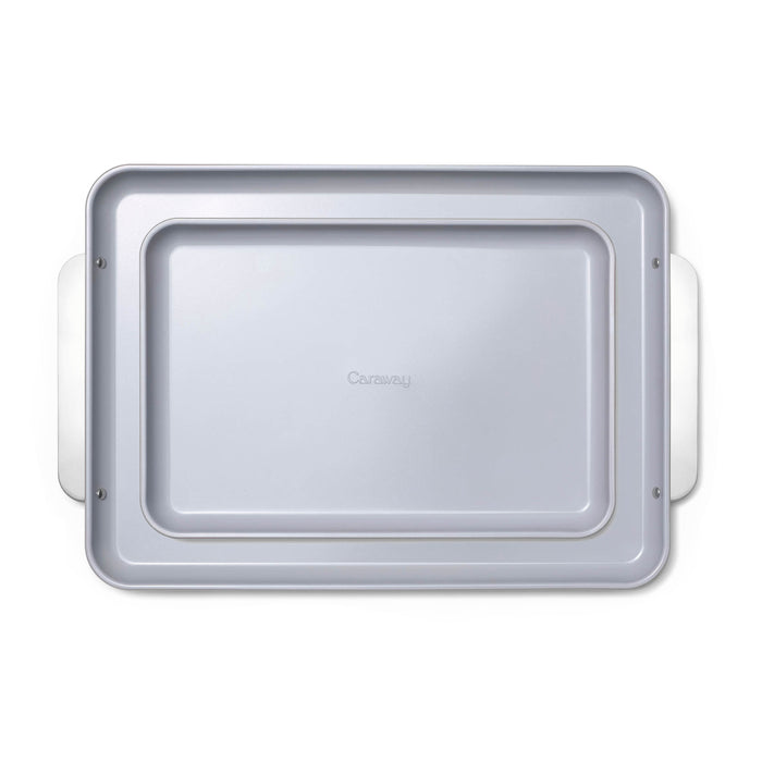 The Caraway Ceramic Baking Sheet Duo is a rectangular metal baking pan with a naturally non-stick, light gray surface and side handles. It features the Caraway brand in the center and is perfect for healthy baking, displayed on a plain white background.
