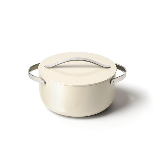 The Caraway Ceramic Dutch Oven, 6.5 qt, is cream-colored with a ceramic-coated finish and includes a matching lid. It features a naturally non-stick surface and metal handles, boasting a sleek modern design perfect for light surfaces.