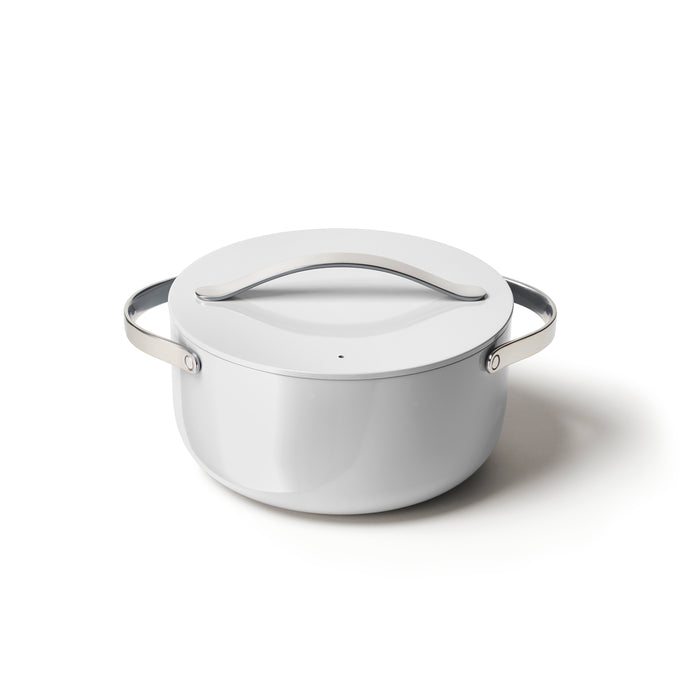 A sleek, white Caraway Ceramic Dutch Oven (6.5 qt) with a lid and polished stainless steel handles sits on a plain white background. Its non-toxic modern design features a smooth surface and a small steam hole on the lid.